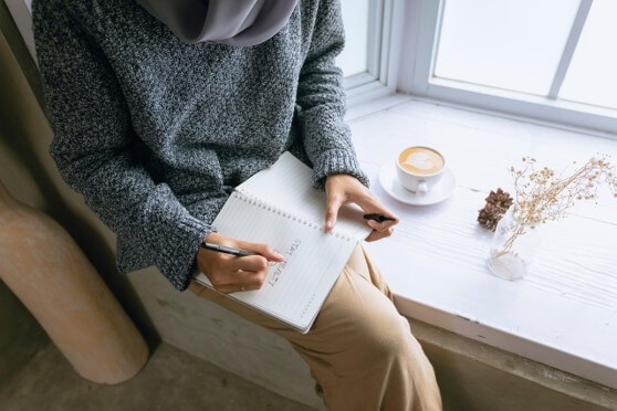 Professional woman in Texas, in her 30s sitting quietly with a notebook after work, reflecting as the sun sets through an office window, representing recovery from burnout as a high achiever.